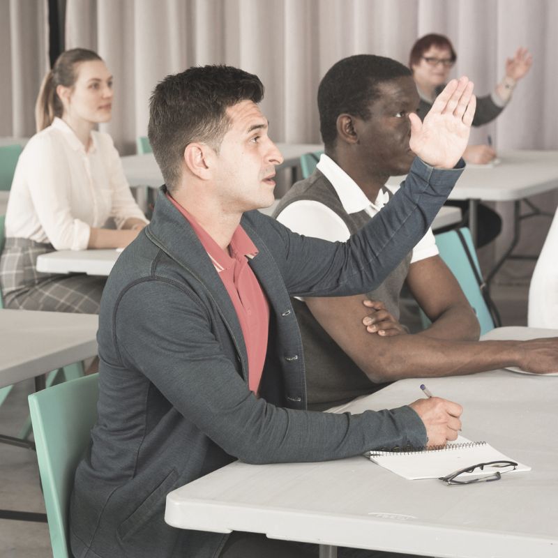 A group of people are in a room sat at desks and many people have their hands up.