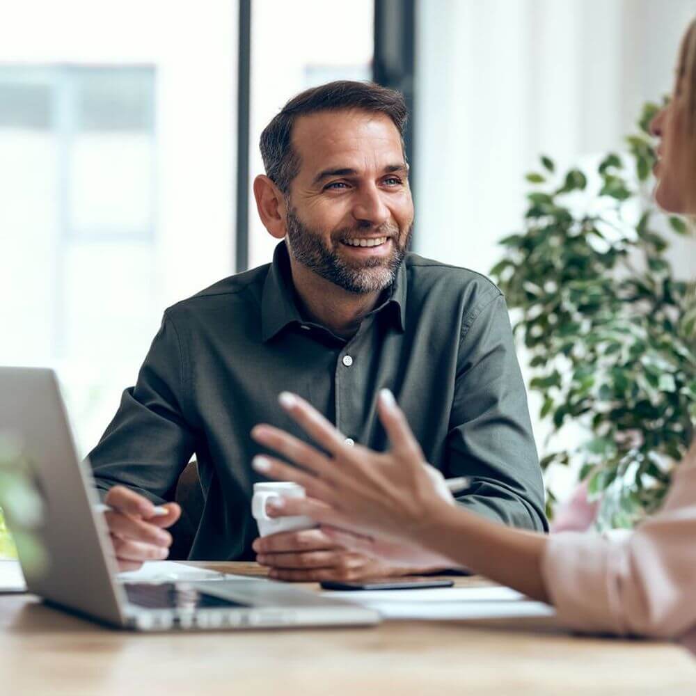 Smiling man holding a coffee cup in conversation with a colleague at an office desk with a laptop open.